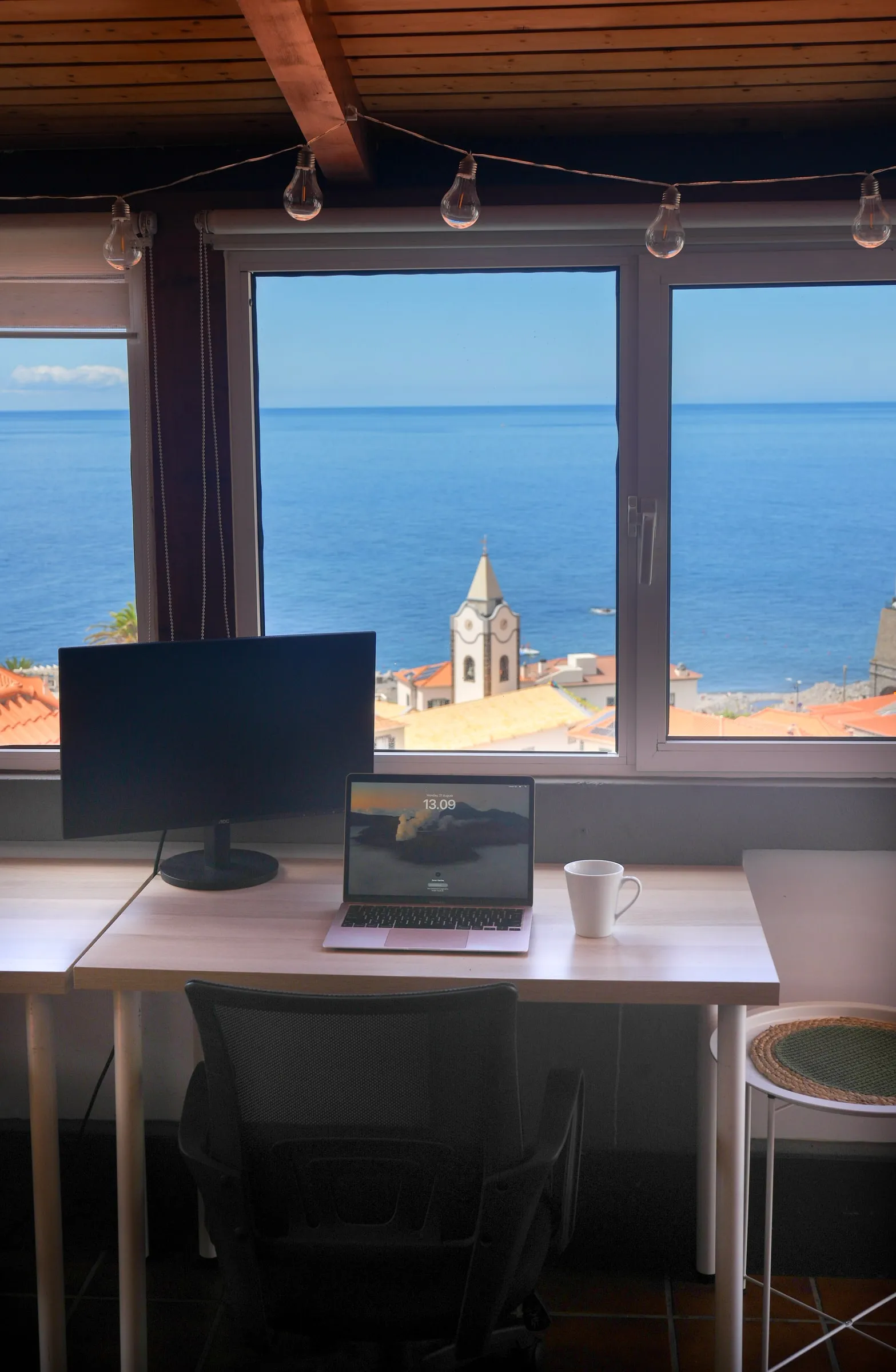 A shared desk and monitor with the village church and ocean beyond the window